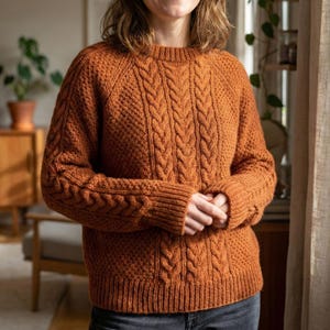 May include: A rust-colored, cable-knit sweater with a crew neck. The sweater features intricate cable patterns down the front and on the sleeves. The knitwear is a warm, cozy style, perfect for cooler weather.
