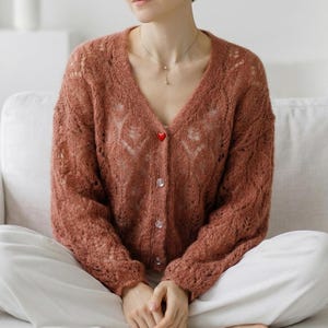 May include: A rust-colored, open-weave cardigan with a V-neck and long sleeves. The cardigan features a heart-shaped red button at the top and several clear buttons down the front. The garment is worn over white pants.