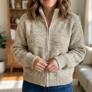 Könnte beinhalten: Beige Reißverschluss-Strickjacke mit Sprenkelmuster. Die Strickjacke hat einen hohen Kragen und gerippte Bündchen und Saum. Der Reißverschluss ist silberfarben. Das Kleidungsstück besteht aus dickem, strukturiertem Strick.