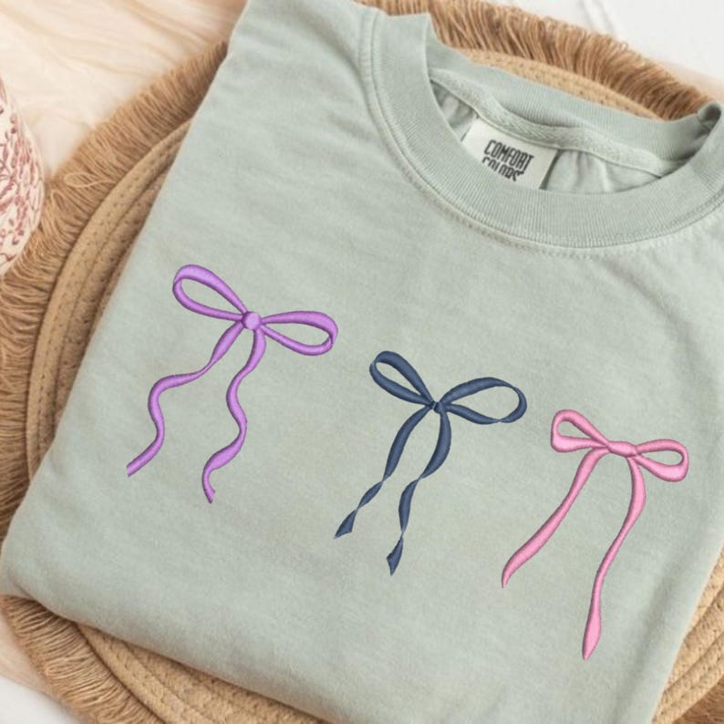 May include: A sage green crewneck t-shirt featuring three embroidered bows in purple, navy blue, and light pink. The shirt has a Comfort Colors brand label inside the collar and a ribbed neckline, showcasing a simple and clean design.