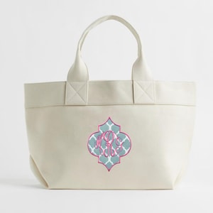May include: Cream-colored tote bag with a decorative quatrefoil design in teal, pink, and white. The design features a monogram in pink. The bag has two handles and is made of a sturdy material.