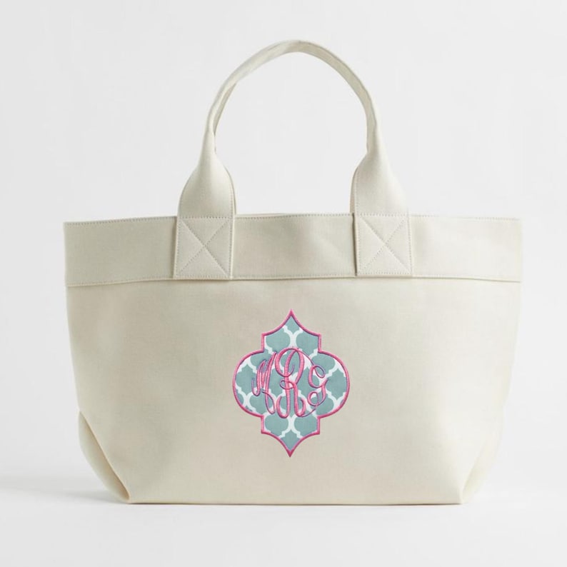 May include: Cream-colored tote bag with a decorative quatrefoil design in teal, pink, and white. The design features a monogram in pink. The bag has two handles and is made of a sturdy material.