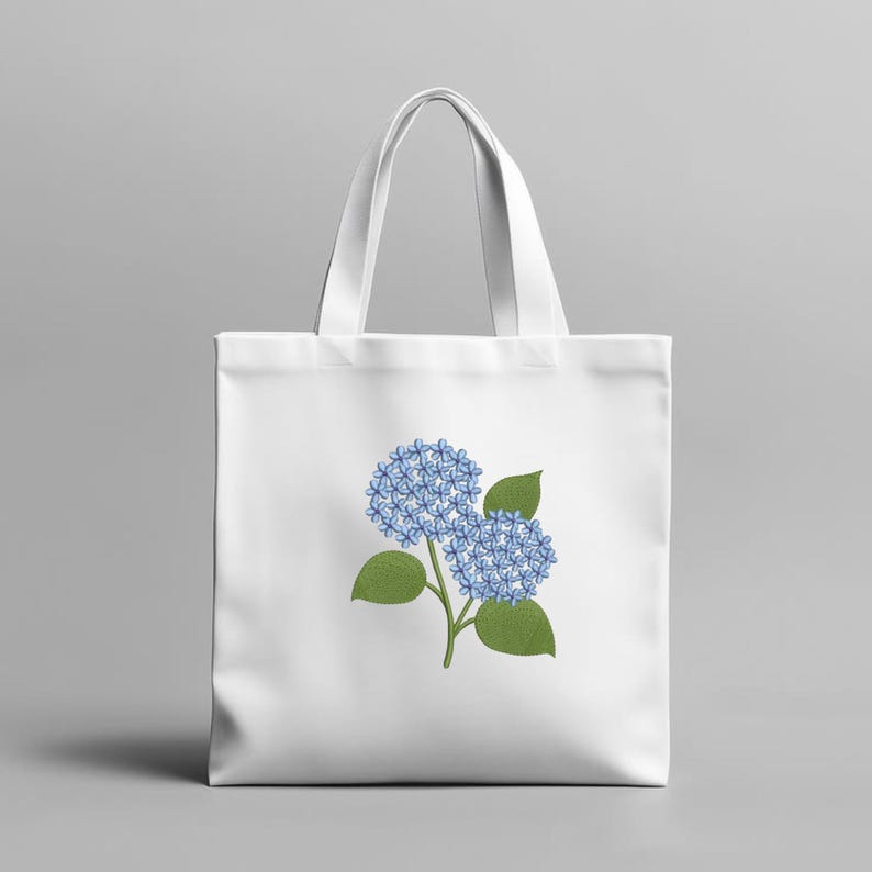 May include: White tote bag featuring a blue hydrangea flower design. The illustration shows two clusters of blue flowers with green leaves and stems. The bag has a simple, clean design, perfect for carrying groceries or personal items.