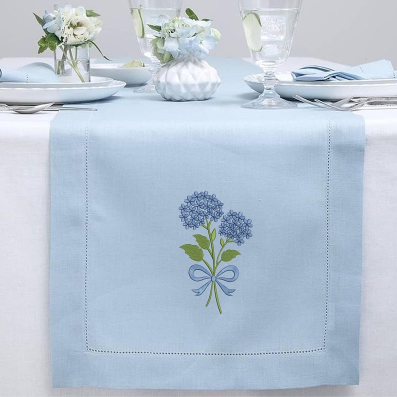 May include: A light blue table runner with a floral hydrangea design. The runner features a stitched border and a detailed illustration of blue hydrangeas with green leaves and a blue bow. The table setting includes white plates, glasses, and small vases with flowers.
