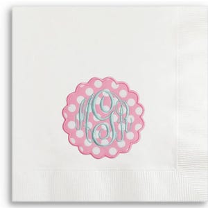 May include: White linen napkin with a scalloped pink and white polka dot emblem. The emblem features a monogram in light blue script. The napkin has a textured border.