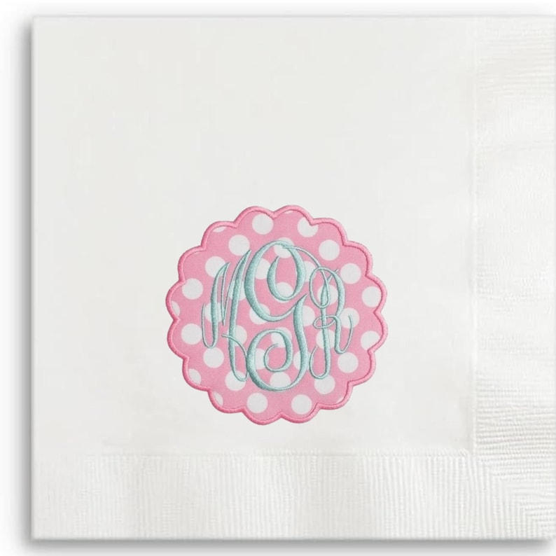 May include: White linen napkin with a scalloped pink and white polka dot emblem. The emblem features a monogram in light blue script. The napkin has a textured border.