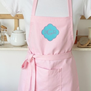 May include: A light pink apron with a large front pocket and a matching waist tie. The apron features a turquoise and pink monogram design on the chest. The apron is worn over a white shirt.
