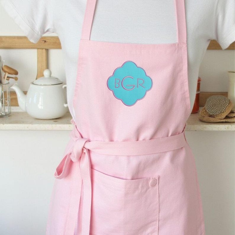 May include: A light pink apron with a large front pocket and a matching waist tie. The apron features a turquoise and pink monogram design on the chest. The apron is worn over a white shirt.