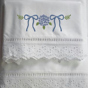 May include: White bedding set with embroidered blue hydrangea and bow design. The top edge features a decorative eyelet trim, and the bottom edge has a scalloped eyelet lace trim. The overall design is elegant and floral.
