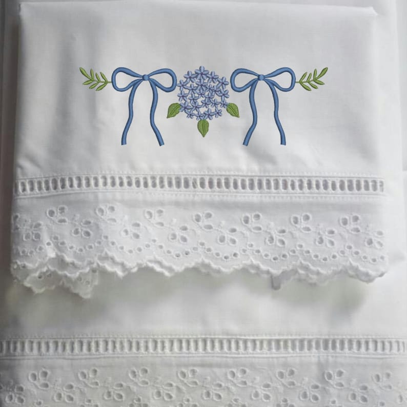 May include: White bedding set with embroidered blue hydrangea and bow design. The top edge features a decorative eyelet trim, and the bottom edge has a scalloped eyelet lace trim. The overall design is elegant and floral.