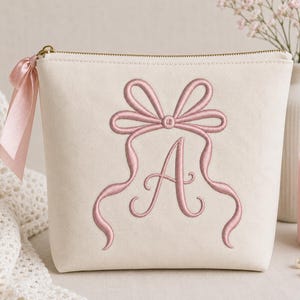 May include: A cream-colored zippered pouch featuring an embroidered dusty pink bow design centered above a large, elegant letter A. A matching dusty pink satin ribbon is tied to the gold-toned zipper pull on the left side of the pouch.