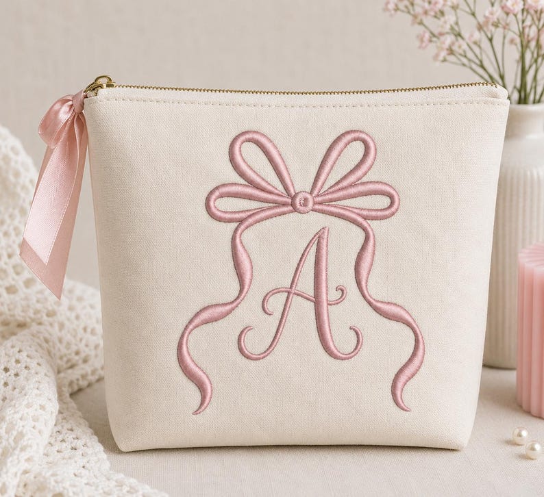 May include: A cream-colored zippered pouch featuring an embroidered dusty pink bow design centered above a large, elegant letter A. A matching dusty pink satin ribbon is tied to the gold-toned zipper pull on the left side of the pouch.