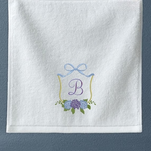 May include: White hand towel with an embroidered design. The design features a light blue bow, a gold-colored shield, the letter "B", and a cluster of blue and purple flowers. The towel is made of a soft, absorbent material.