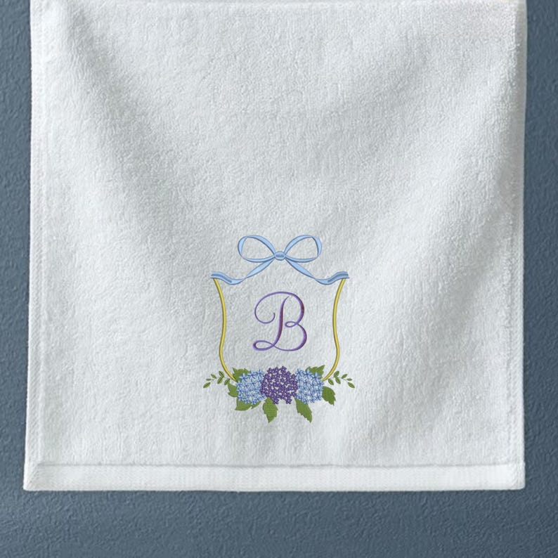 May include: White hand towel with an embroidered design. The design features a light blue bow, a gold-colored shield, the letter "B", and a cluster of blue and purple flowers. The towel is made of a soft, absorbent material.