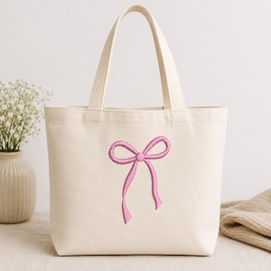 May include: A cream-colored canvas tote bag featuring a centered, embroidered pink ribbon bow design. The bag has two sturdy top handles and a structured rectangular shape, crafted from a woven fabric with a visible texture throughout the material.