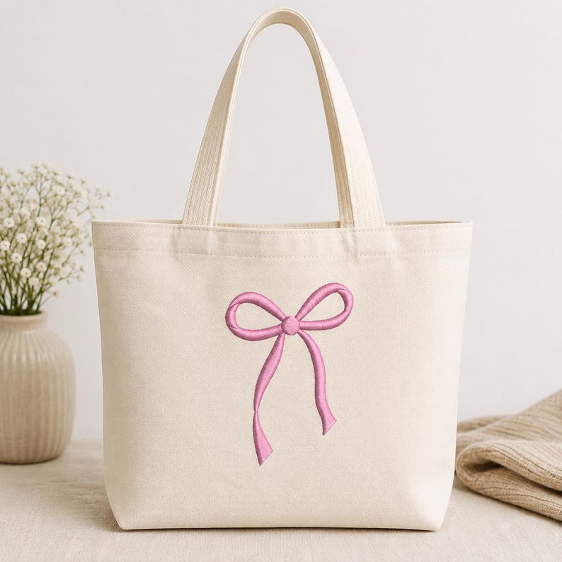 May include: A cream-colored canvas tote bag featuring a centered, embroidered pink ribbon bow design. The bag has two sturdy top handles and a structured rectangular shape, crafted from a woven fabric with a visible texture throughout the material.