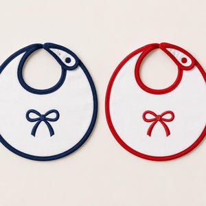 May include: Two round white bibs with a snap closure at the neck. One bib features a dark blue trim and a matching dark blue embroidered bow in the center. The other bib features a red trim and a matching red embroidered bow in the center.