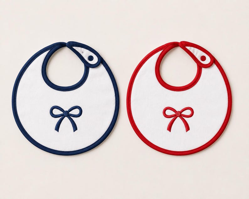 May include: Two round white bibs with a snap closure at the neck. One bib features a dark blue trim and a matching dark blue embroidered bow in the center. The other bib features a red trim and a matching red embroidered bow in the center.