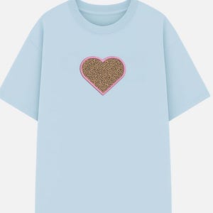 May include: A light blue t-shirt featuring a heart-shaped design. The heart has a leopard print pattern with a pink border. The shirt has short sleeves and a crew neck.