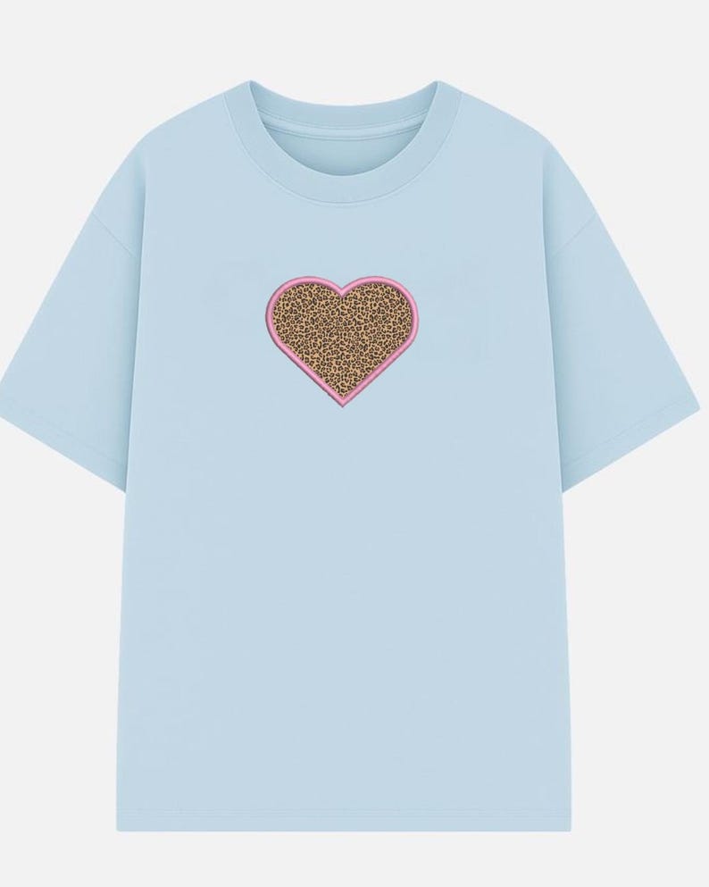 May include: A light blue t-shirt featuring a heart-shaped design. The heart has a leopard print pattern with a pink border. The shirt has short sleeves and a crew neck.