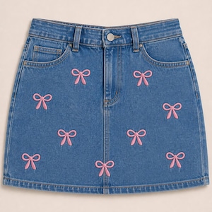 May include: A blue denim mini skirt featuring a repeating pattern of embroidered light pink bows. The skirt includes a standard waistband with belt loops, a metal button closure, a front zipper fly, and two front pockets with visible stitching.