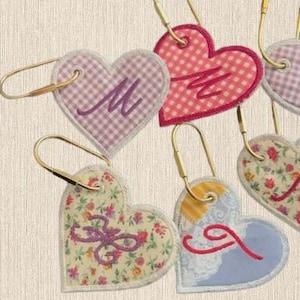 May include: A collection of heart-shaped fabric tags, each attached to a gold-toned metal ring. The tags feature various patterns including pink gingham, floral prints, and blue designs, with embroidered letters M, J, and other stylized monograms.