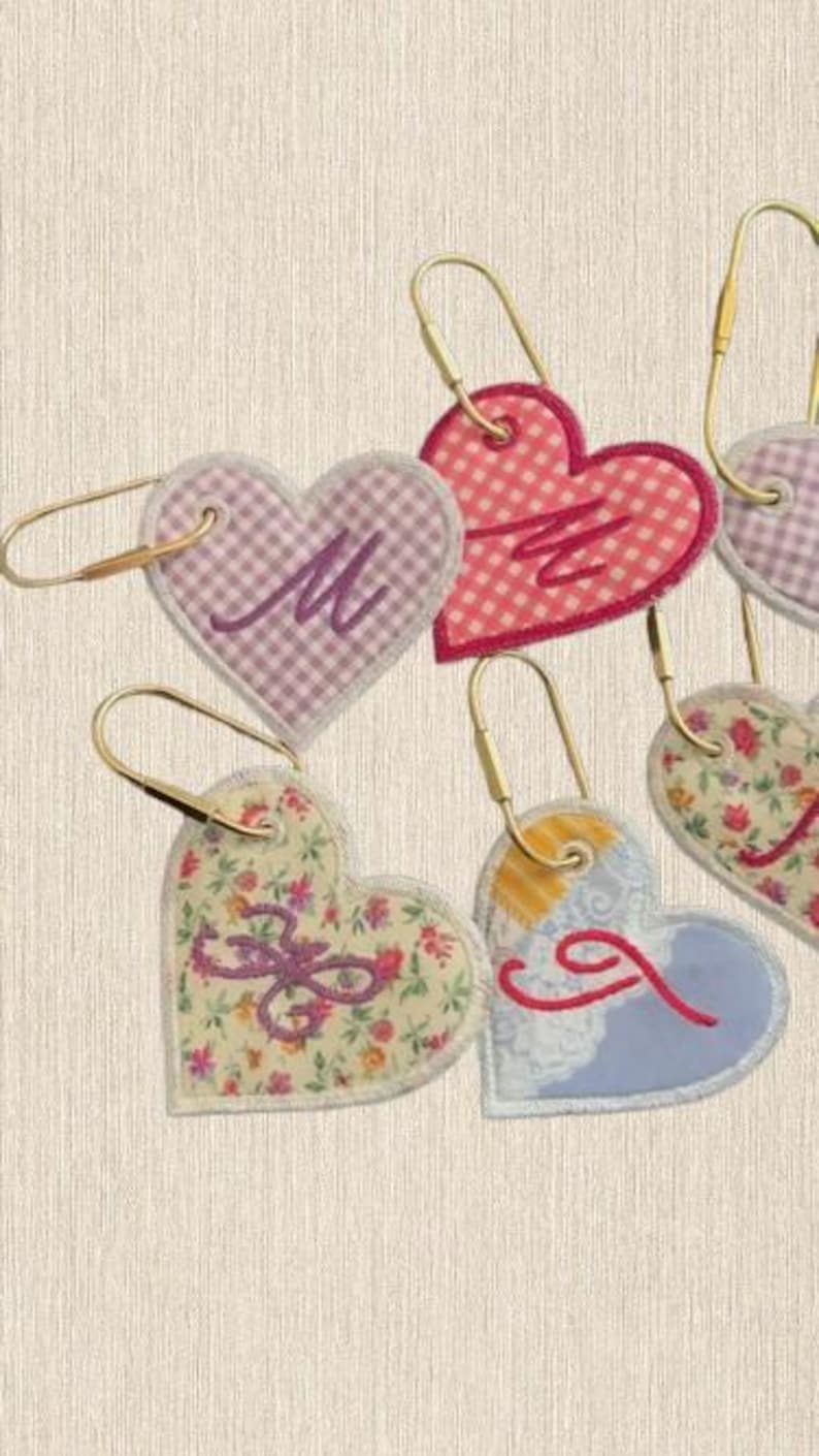 May include: A collection of heart-shaped fabric tags, each attached to a gold-toned metal ring. The tags feature various patterns including pink gingham, floral prints, and blue designs, with embroidered letters M, J, and other stylized monograms.