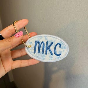 May include: An oval-shaped fabric keychain featuring a light blue and white floral pattern with the letters MKC embroidered in dark blue thread. The fabric patch is attached to a gold-toned metal ring with a screw-style closure mechanism.