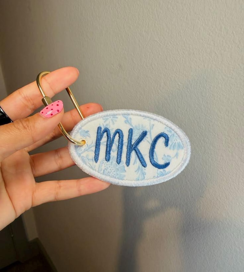 May include: An oval-shaped fabric keychain featuring a light blue and white floral pattern with the letters MKC embroidered in dark blue thread. The fabric patch is attached to a gold-toned metal ring with a screw-style closure mechanism.