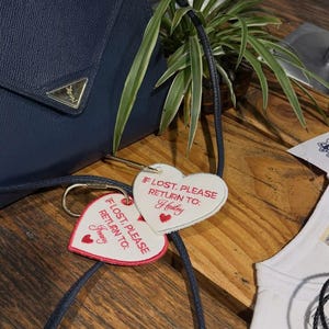May include: A navy blue textured handbag with a gold triangular logo plaque, featuring two white heart-shaped luggage tags attached by gold rings. The tags are embroidered with red text reading IF LOST, PLEASE RETURN TO: followed by the names Hailey or Jenny.