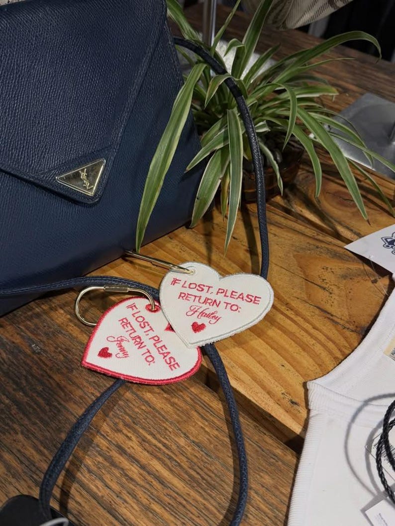 May include: A navy blue textured handbag with a gold triangular logo plaque, featuring two white heart-shaped luggage tags attached by gold rings. The tags are embroidered with red text reading IF LOST, PLEASE RETURN TO: followed by the names Hailey or Jenny.