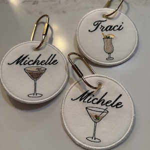 May include: Three circular white fabric tags with gold-toned metal lobster clasps. Each tag features black embroidered names: Michelle, Traci, and Michele. Below each name is a small embroidered cocktail glass with a garnish.