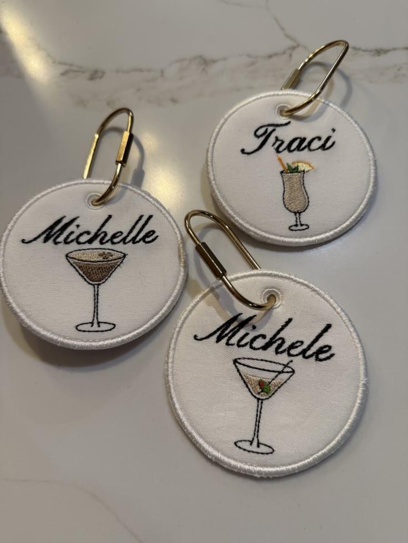 May include: Three circular white fabric tags with gold-toned metal lobster clasps. Each tag features black embroidered names: Michelle, Traci, and Michele. Below each name is a small embroidered cocktail glass with a garnish.