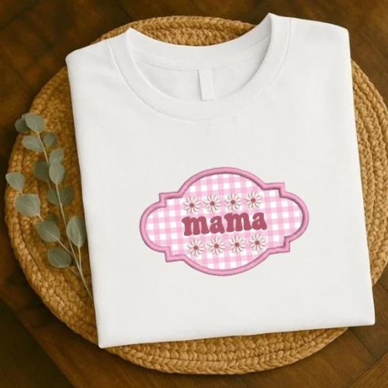 May include: White t-shirt with a pink and white gingham design that says "mama". The design is framed by a pink border and decorated with small white flowers. The shirt is displayed on a woven mat.