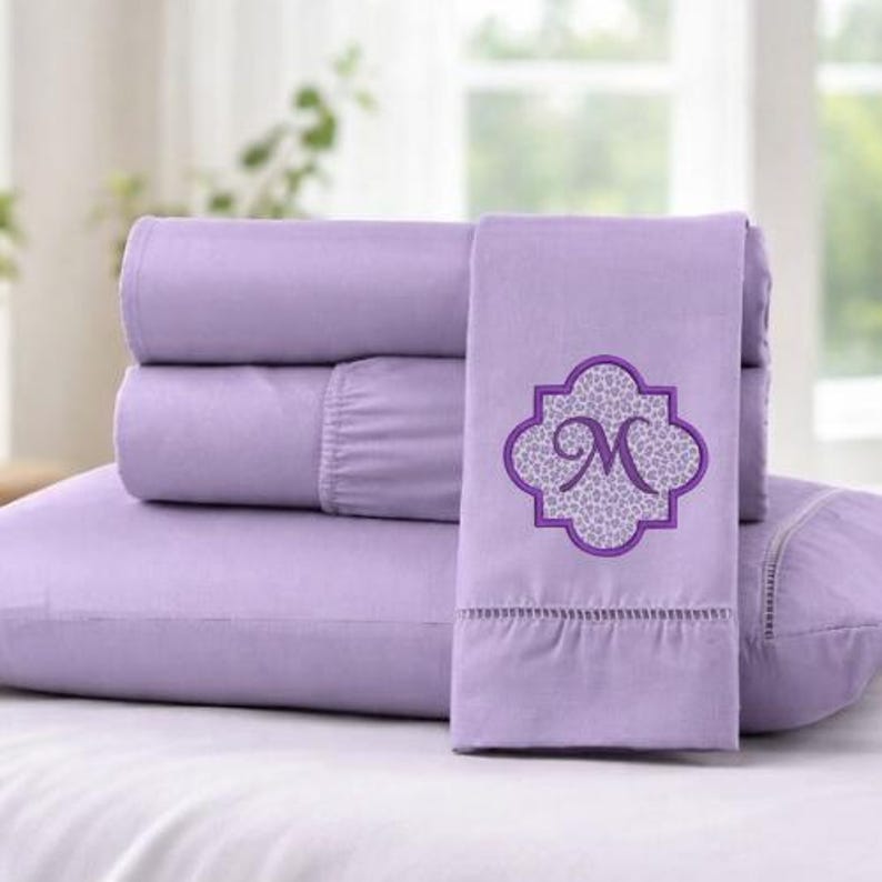 May include: A lavender-colored sheet set with an embroidered design. The set includes a fitted sheet, flat sheet, and pillowcases. The embroidered design is a purple quatrefoil shape with a leopard print background and the letter "M".