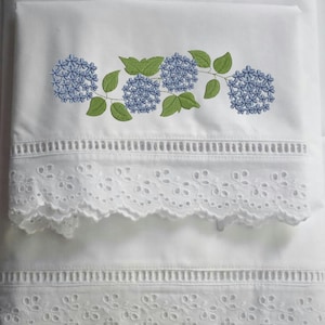 May include: White bedding with embroidered blue hydrangea flowers and green leaves. The bedding features a decorative eyelet lace trim along the edges, adding a delicate touch to the design. The overall aesthetic is fresh and floral.