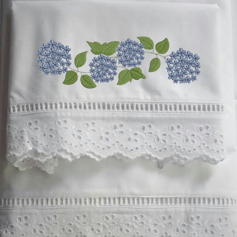 May include: White bedding with embroidered blue hydrangea flowers and green leaves. The bedding features a decorative eyelet lace trim along the edges, adding a delicate touch to the design. The overall aesthetic is fresh and floral.