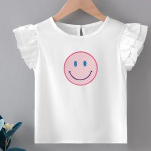 May include: White ruffled-sleeve top with a pink and blue smiley face design. The smiley face has a pink checkered pattern, blue eyes, and a curved smile. The top is hanging on a wooden hanger.