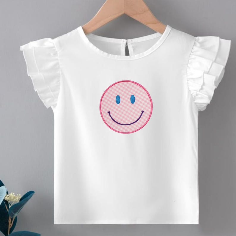 May include: White ruffled-sleeve top with a pink and blue smiley face design. The smiley face has a pink checkered pattern, blue eyes, and a curved smile. The top is hanging on a wooden hanger.