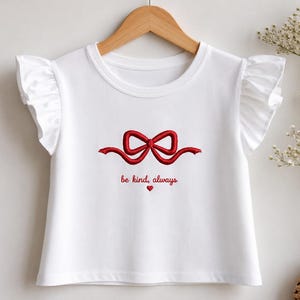 May include: A white short-sleeved t-shirt with ruffled sleeves and a crew neckline. The center features an embroidered red ribbon bow graphic above the red text "be kind, always" and a small red heart icon centered below the text.