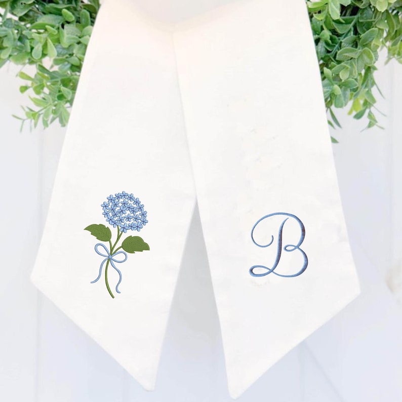 May include: Two white decorative towels. One towel features a blue hydrangea flower with green leaves and a blue ribbon. The other towel has a blue embroidered letter "B". The towels are hanging against a white background.