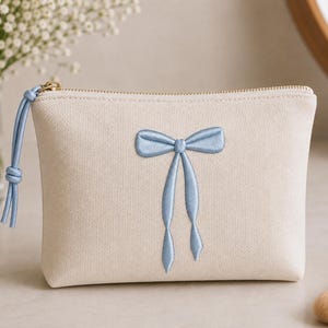 May include: A cream-colored canvas pouch featuring a centered light blue embroidered bow design. The pouch has a gold-toned metal zipper closure with a matching light blue cord pull tab attached to the zipper slider on the left side.