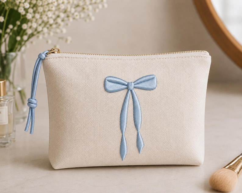 May include: A cream-colored canvas pouch featuring a centered light blue embroidered bow design. The pouch has a gold-toned metal zipper closure with a matching light blue cord pull tab attached to the zipper slider on the left side.