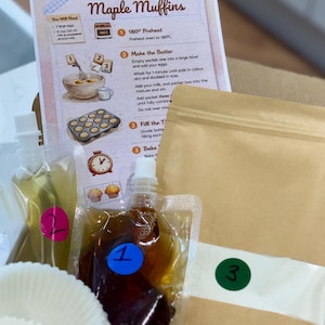 May include: A maple muffin baking kit, containing a recipe card with instructions and illustrations. The kit includes numbered ingredient packets, a muffin liner, and colourful packing materials. The recipe card says "Maple Muffins".