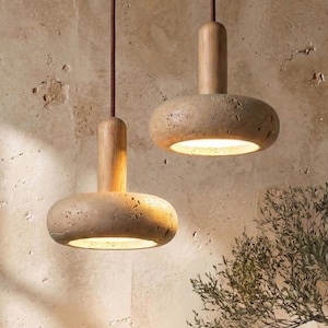 Peut inclure: Deux suspensions lumineuses avec des abat-jours en pierre naturelle et des sommets en bois. Les lampes sont suspendues à des cordons bruns sur un mur texturé brun clair. Les lampes émettent une lueur chaleureuse, créant une ambiance chaleureuse. Design minimaliste et moderne.