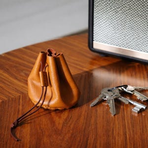 Könnte beinhalten: Ein kleiner, hellbrauner Lederbeutel mit Kordelverschluss liegt auf einer Holzoberfläche neben einem Schlüsselbund. Im Hintergrund befindet sich ein Lautsprecher.