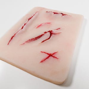 Puede incluir: Un cojín rectangular de silicona color carne con varias laceraciones simuladas. Los cortes son de varias longitudes y profundidades, llenos de pigmento rojo para imitar la sangre. El cojín está diseñado para entrenamiento médico o de efectos especiales.