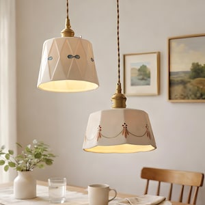 Puede incluir: Dos lámparas colgantes con pantallas de color crema cuelgan sobre una mesa de comedor. Una pantalla tiene un patrón geométrico con acentos azules, la otra tiene un diseño floral. Las luces tienen herrajes de color latón y están suspendidas por cables marrones.
