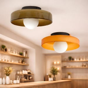 May include: Two modern ceiling lights with a round, flat design. One light has a dark olive green shade, the other is orange. Both have a white globe light in the center and a black top. The lights are in a kitchen or dining area.