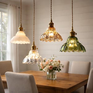 Può includere: Quattro lampade a sospensione con paralumi in vetro smerlato bianco, ambra, trasparente e verde sono appese sopra un tavolo da pranzo in legno. Ogni lampada ha un apparecchio in ottone e un cavo intrecciato. Un vaso di fiori è sul tavolo.